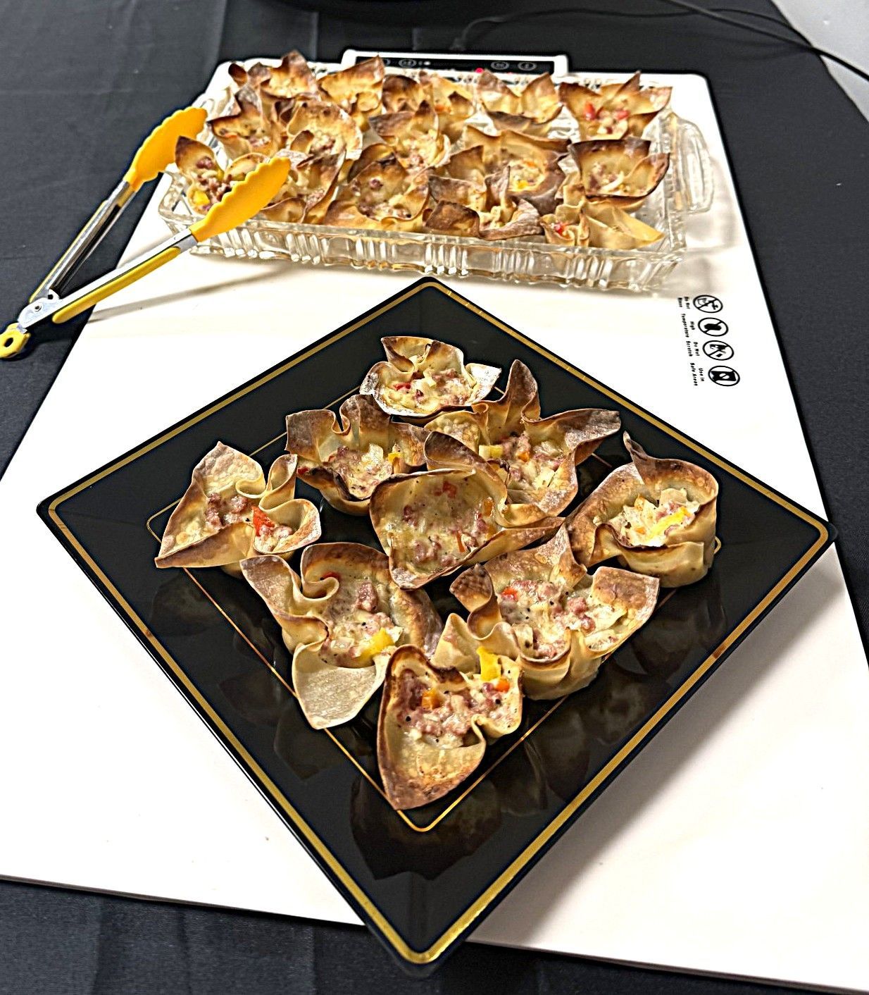 Two plates of baked flower-shaped pastries on a black and a foil tray, with tongs on the side
