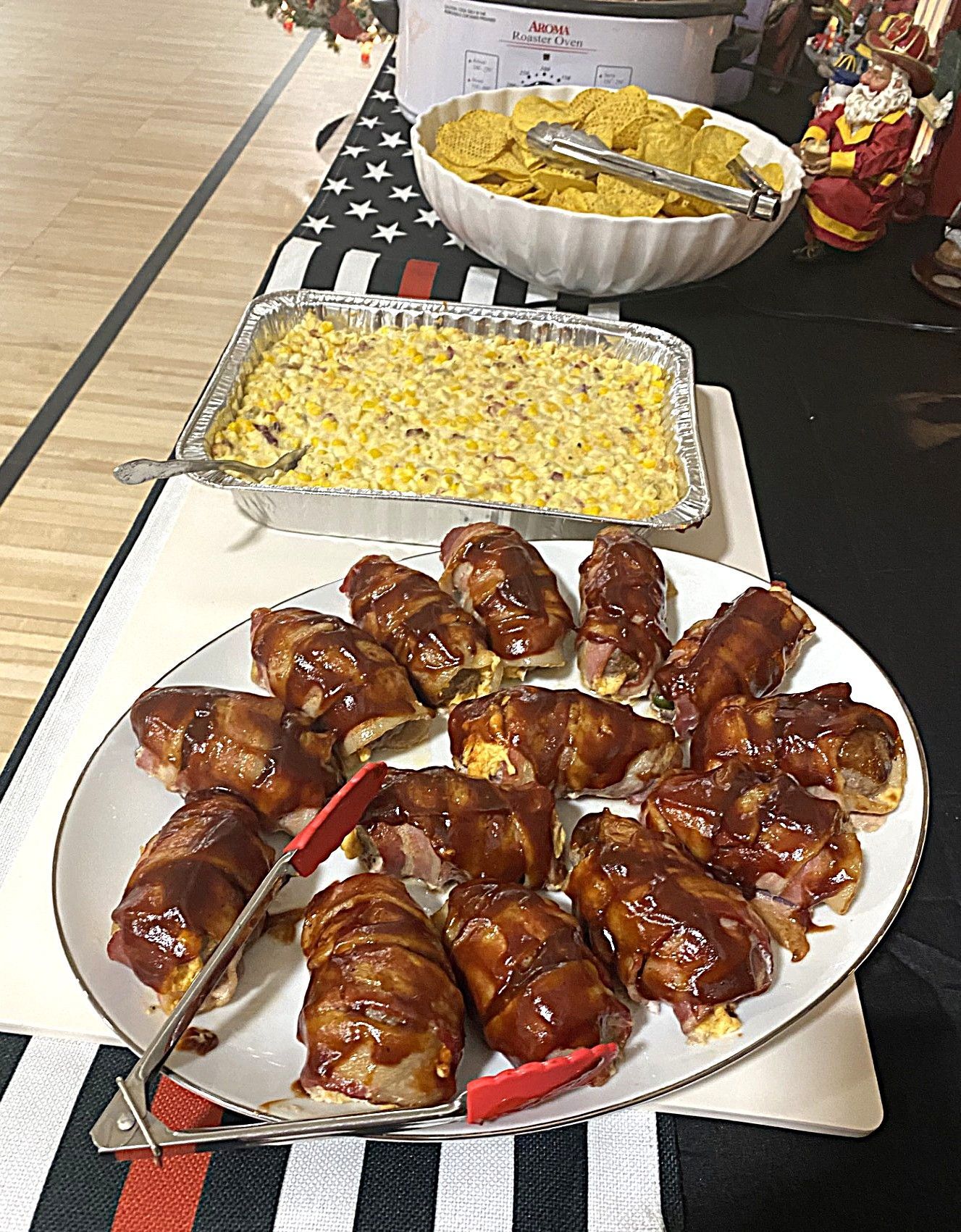 Tray of glazed bacon-wrapped appetizers on a buffet table, with a casserole of cheesy pasta and chips behind.