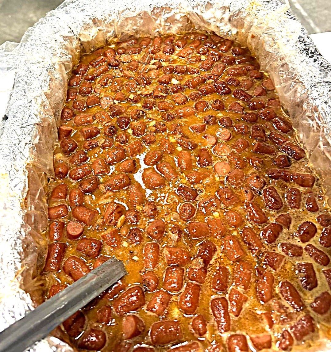 Tray of baked beans with a spoon, covered in shiny sauce and foil-lined edges.