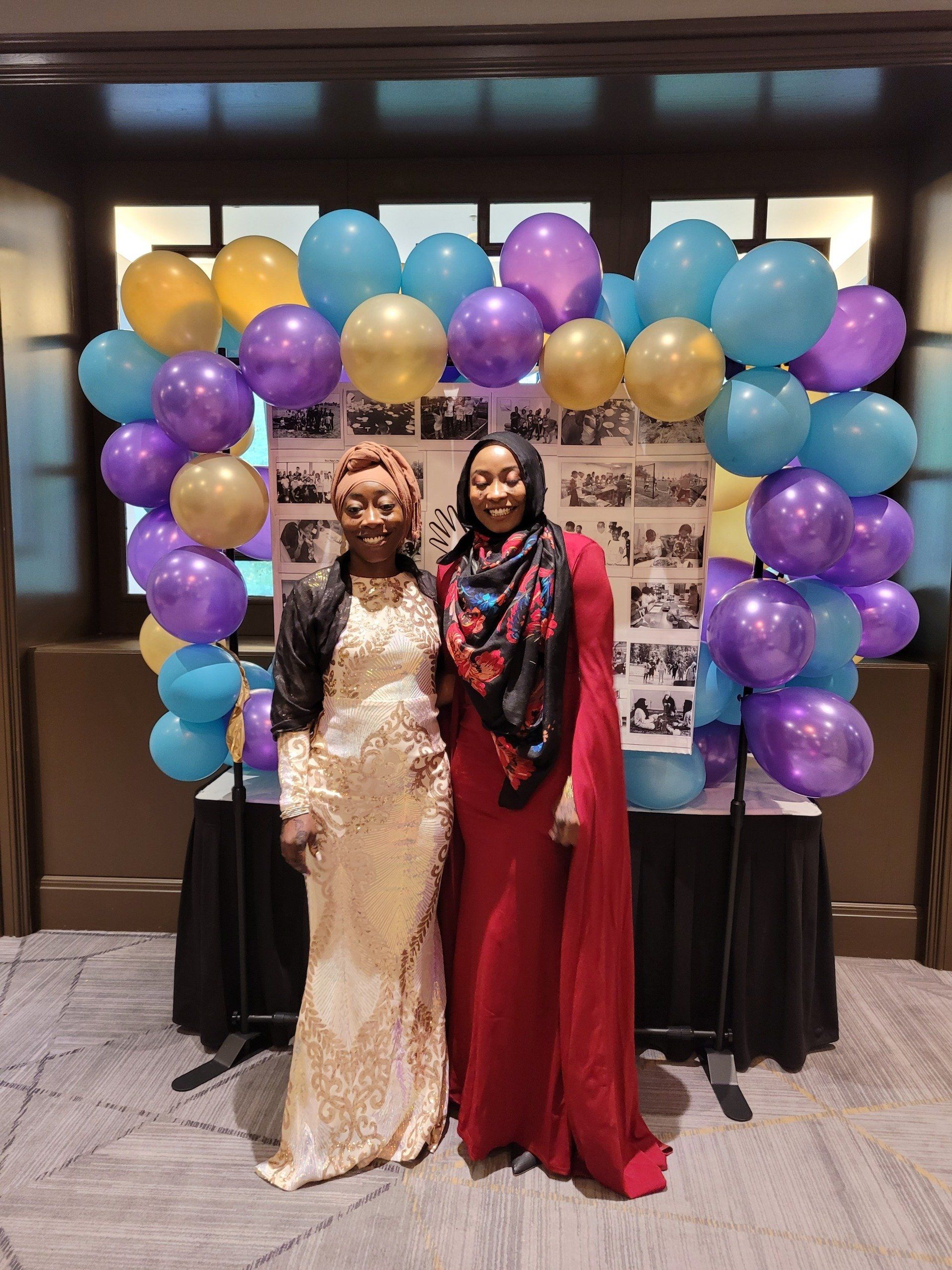 Two women are posing for a picture in front of a wall of balloons.