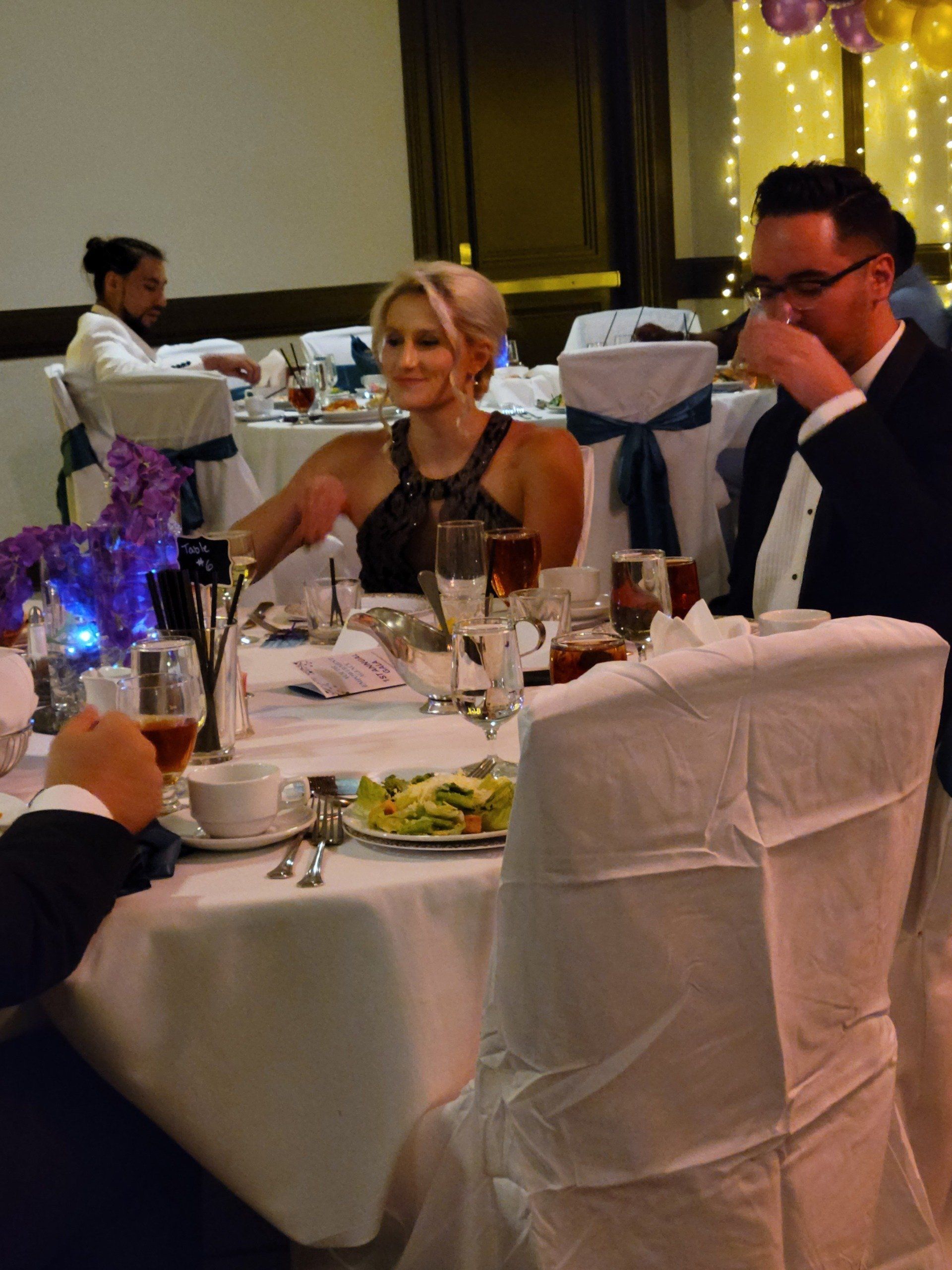 A woman in a black top sits at a table with a man in a tuxedo