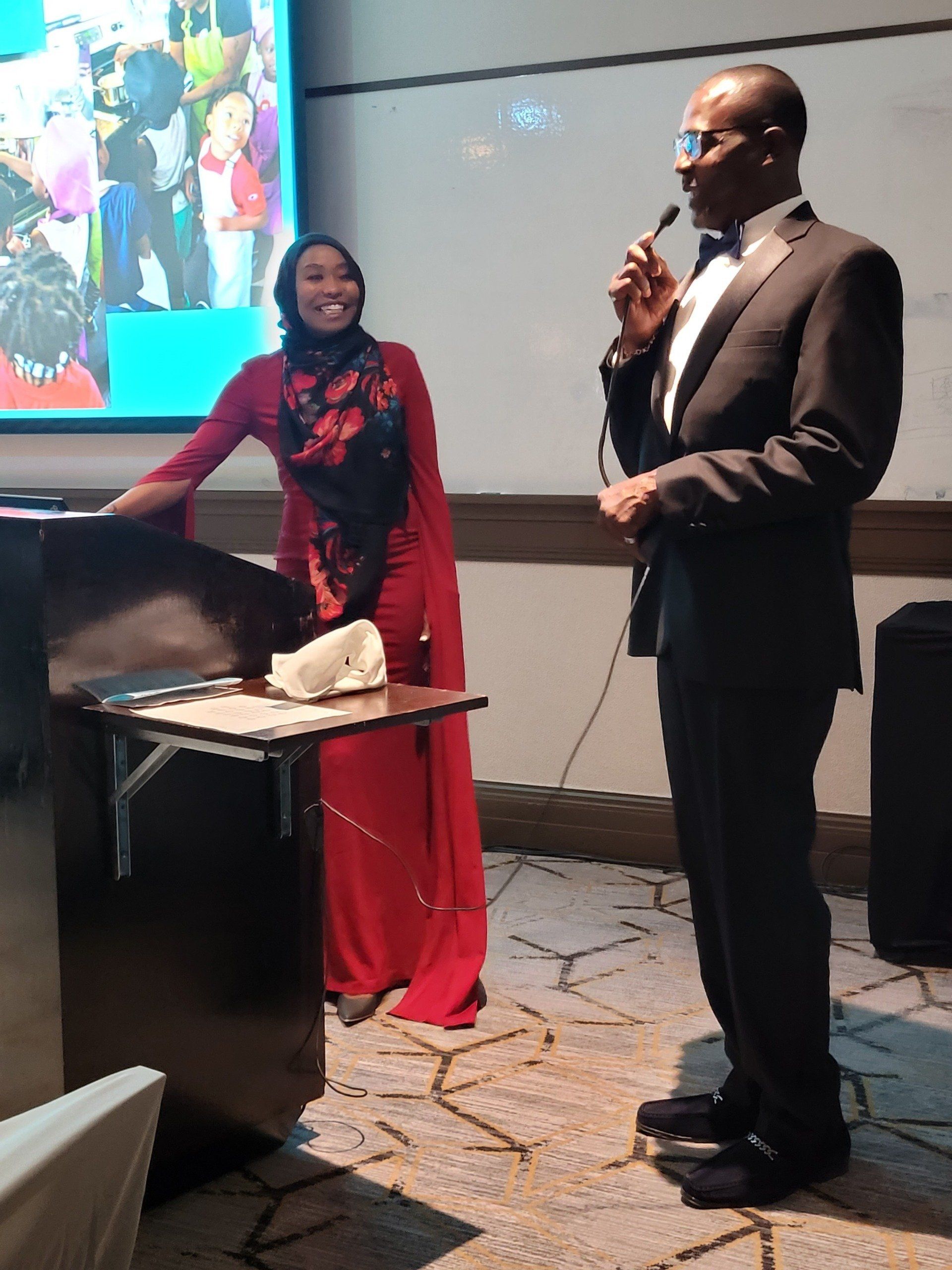 A man in a tuxedo is speaking into a microphone while a woman in a red dress stands behind him