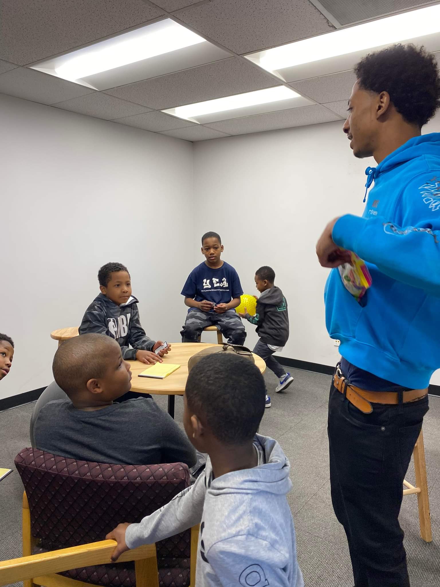 A man in a blue hoodie is standing in front of a group of children sitting around a table.