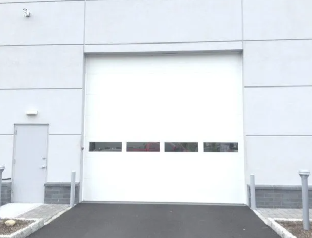 White industrial building with a large garage door and a small side door on a paved driveway.