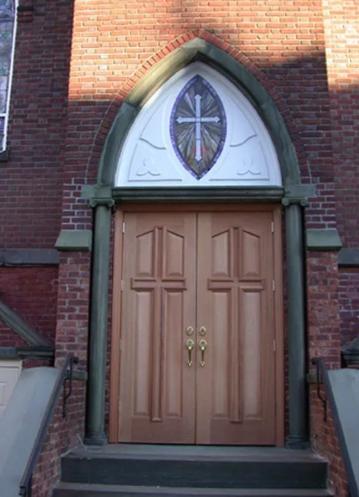 Wooden double church doors beneath a pointed arch and cross window in a brick wall