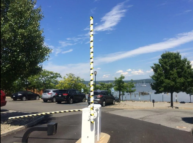 Parking lot by waterfront with striped barrier pole, cars, trees, and benches under a blue sky.