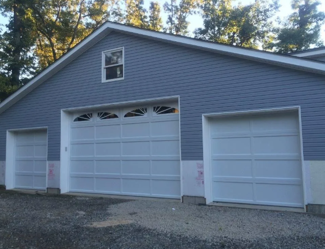 Gray garage with three white doors and a small window under a gabled roof, set by trees and gravel driveway