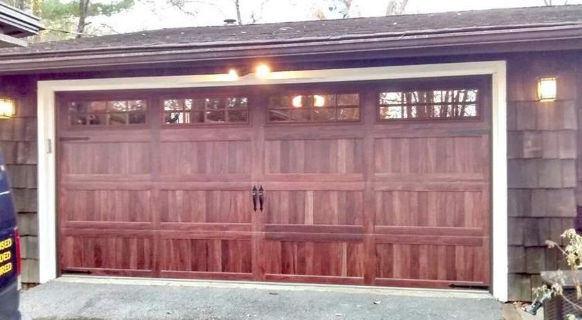 Brown double garage door on a house exterior at dusk, with white trim and lights on above it