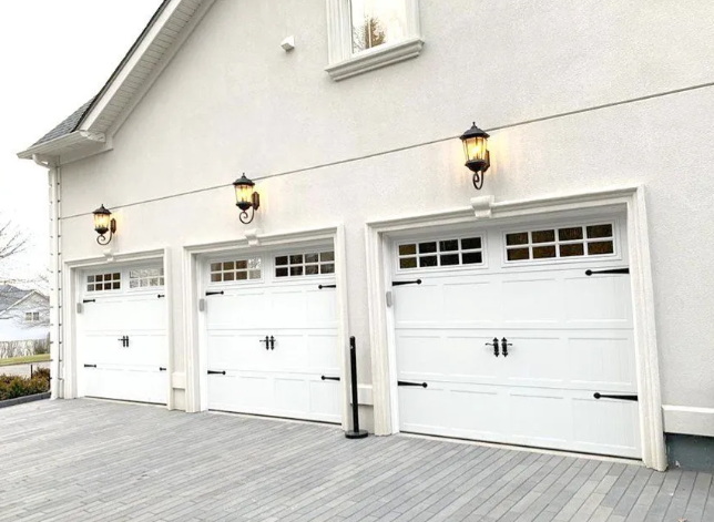 Three white garage doors on a light gray house with outdoor lanterns and a wooden deck