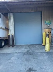 Industrial loading dock with a closed blue roll-up door and yellow bollards at the entrance