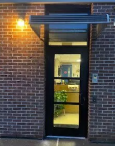 Brick building entrance with lit wall lamp and a glass door showing a lobby inside