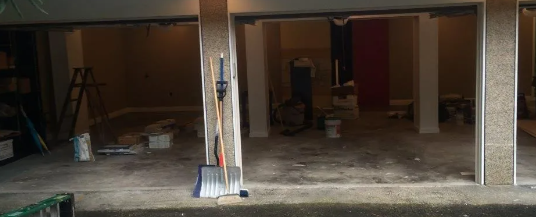 Empty storefront with open garage bays, wet concrete floor, and brooms leaning by the entrance.