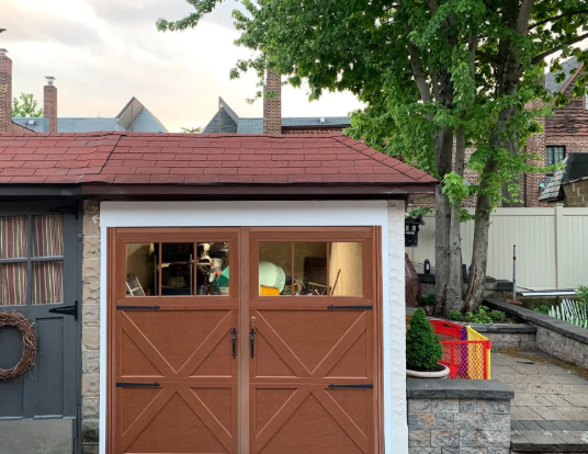 Brown garage with white trim in a backyard, with a tree and patio area beside it