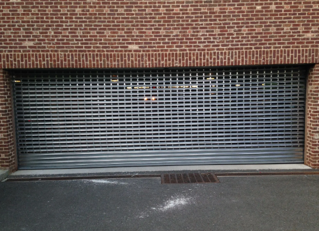 Closed metal security shutter on a brick building, with a drain grate on the sidewalk.