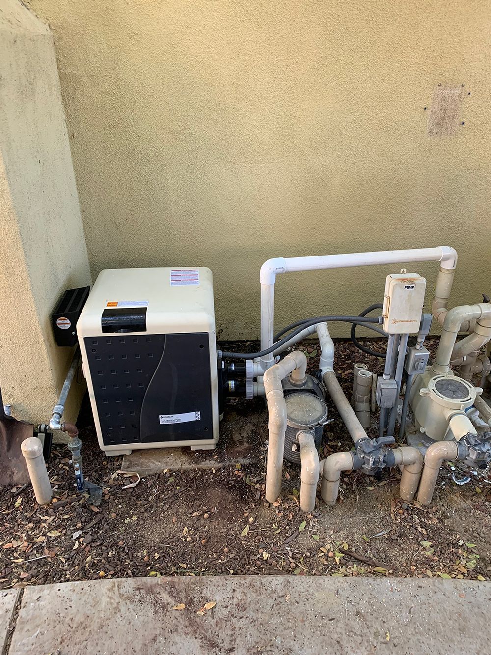 Pool equipment including a heater, pipes, and filter against a beige wall.