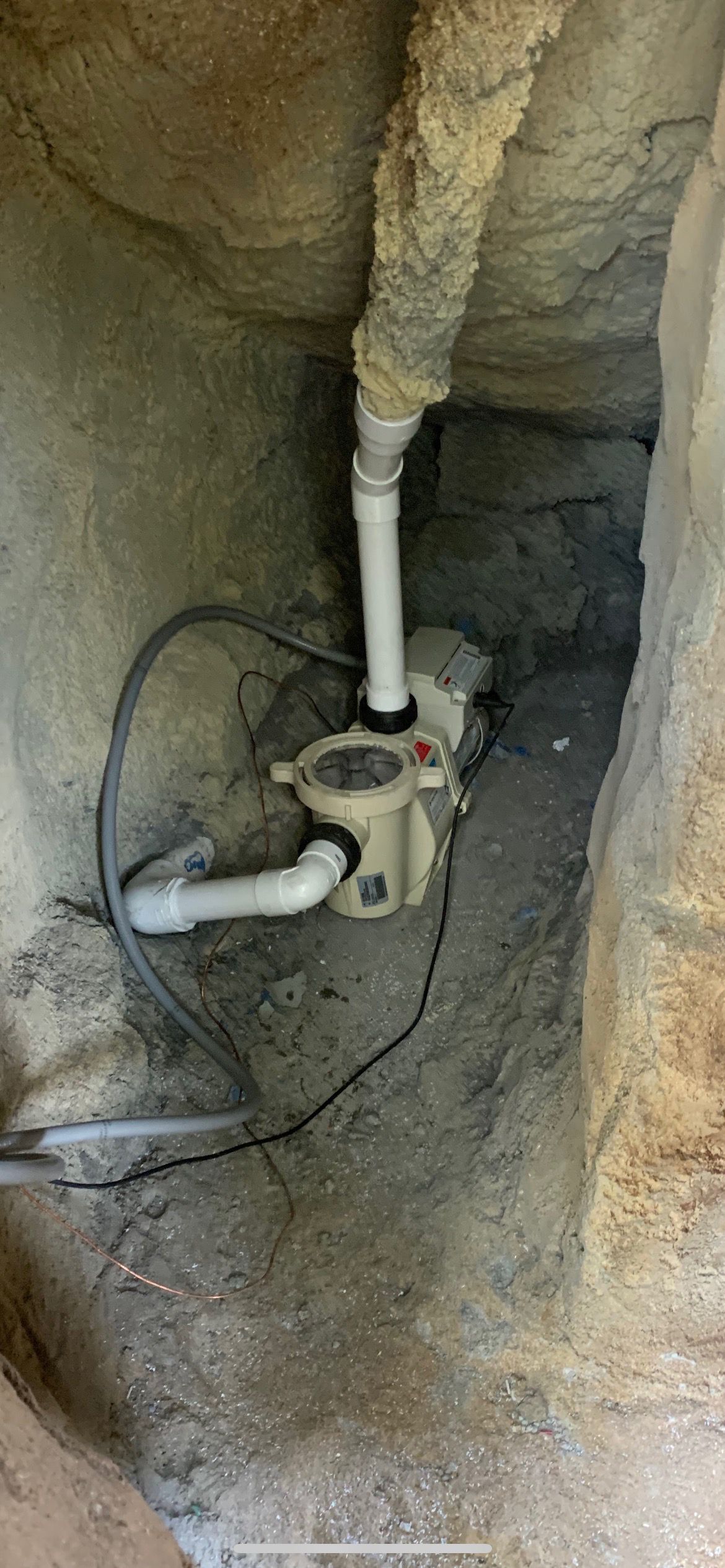 Pool pump in a rocky alcove. White pipes and black electrical cable.