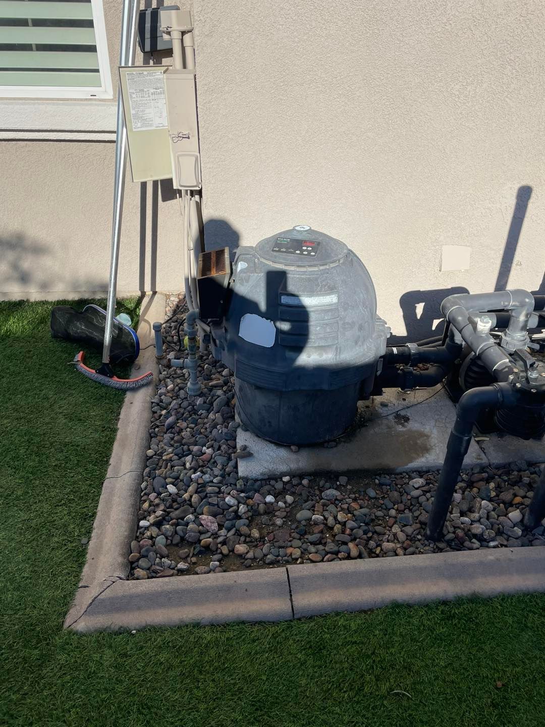 Pool filter equipment next to a beige wall. Equipment sits on a concrete base surrounded by gravel.