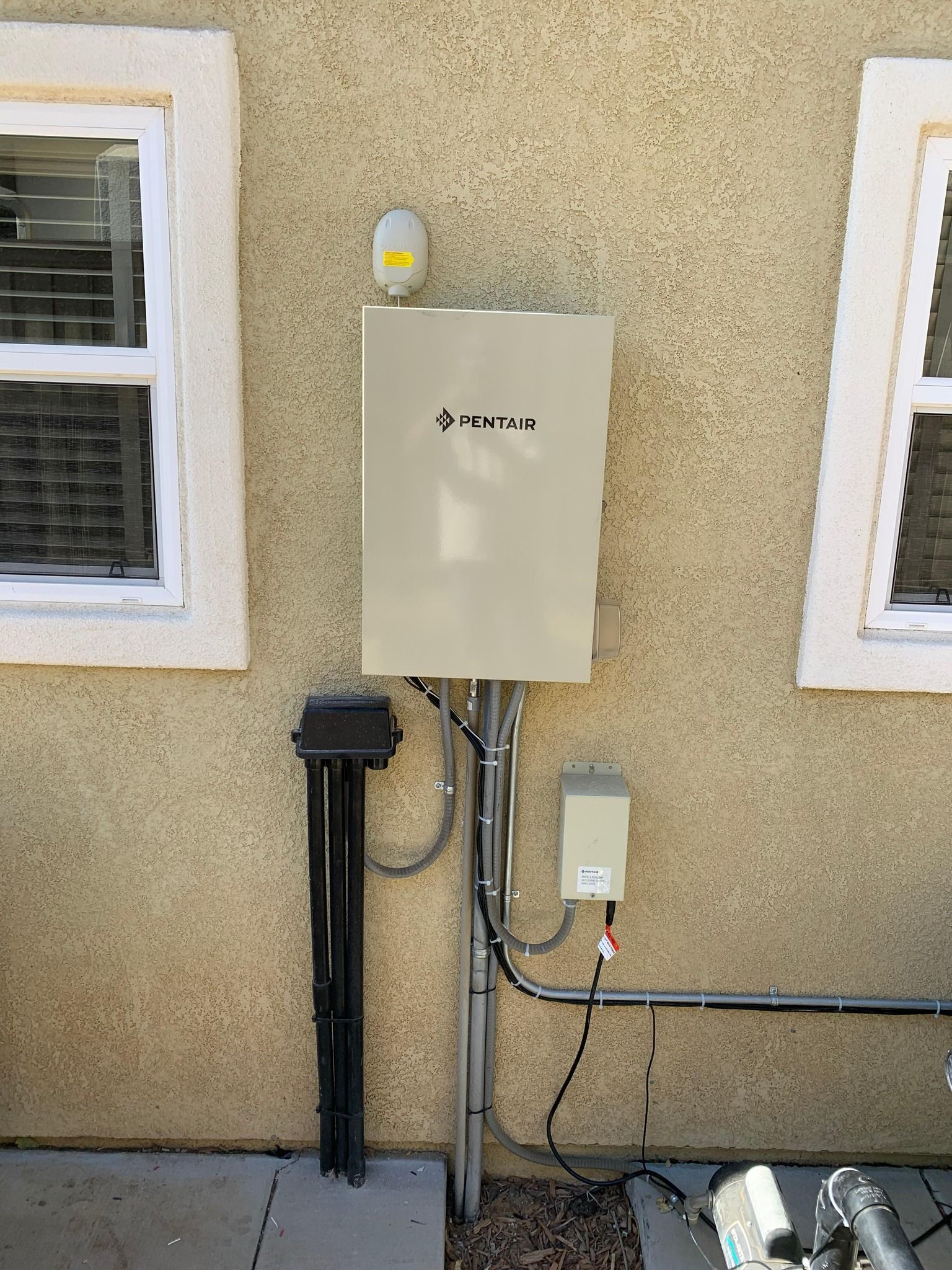 Beige utility box on tan stucco wall, flanked by windows. Cables and antenna visible.