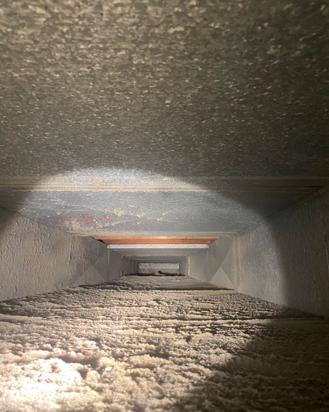 Dust-filled interior of an HVAC duct, viewed from within. Gray metal walls and a layer of white dust are visible.
