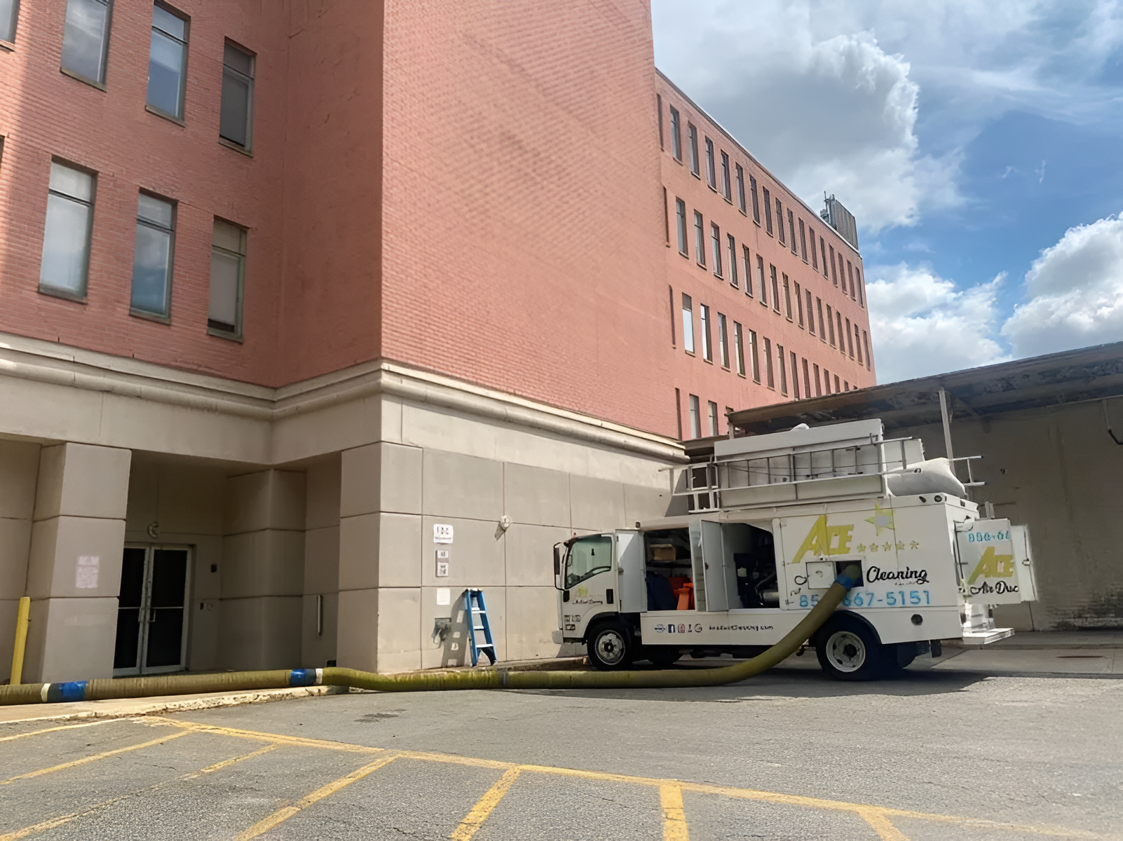 A service truck connected to a building with a hose, possibly cleaning. Red brick building, parking lot.