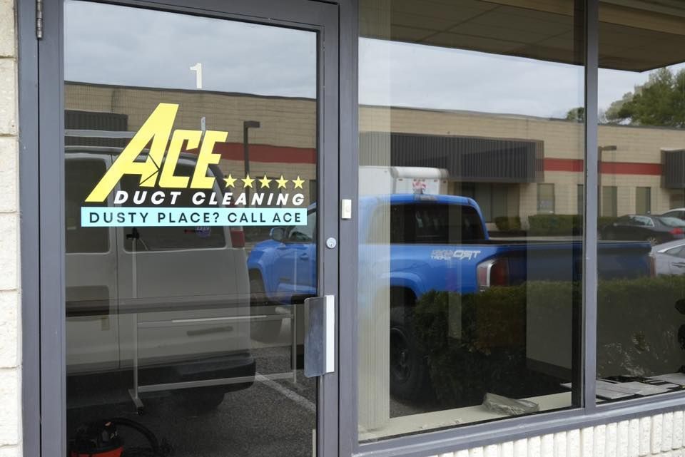 Ace Duct Cleaning storefront with logo on glass door, building and vehicles reflected in window.