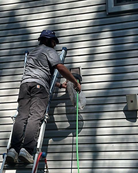 Person on a ladder cleaning a vent on the side of a house. They are holding a green hose.