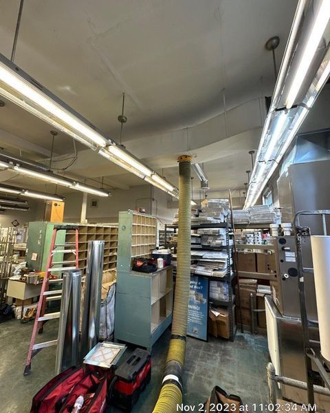 Inside a cluttered workshop: storage shelves, tools, equipment, overhead lights, ventilation hose.