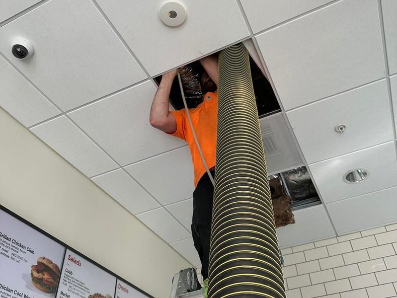 Man in orange shirt working in ceiling tiles, vacuum hose extended.