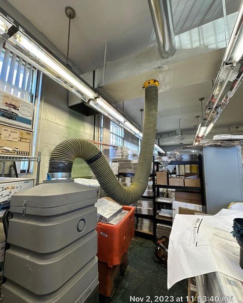 Gray ventilation hose suspended from the ceiling in a cluttered room with shelves and equipment.