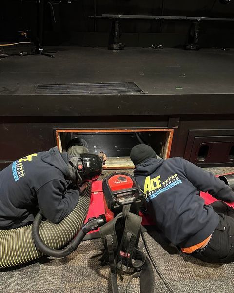 Two workers in blue hoodies and masks are vacuuming a dark opening, near a stage.