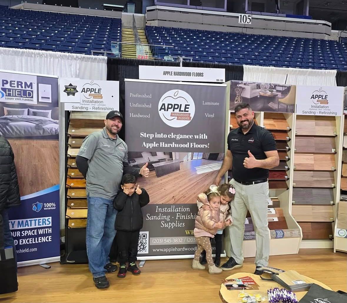 Two men posing with an Apple Mattress display at an indoor stadium booth, with stacked mattresses and product signs.