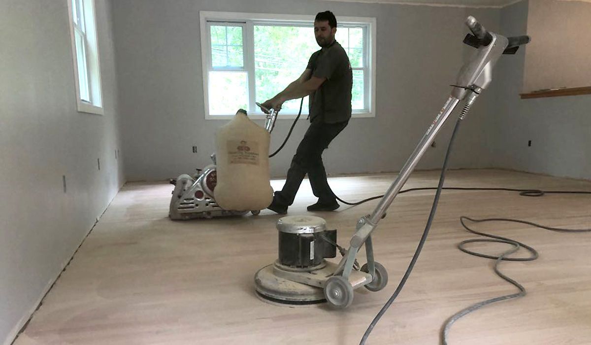 Worker sanding a floor with a floor sander in an empty room, with a vacuum hose and extension pole nearby.
