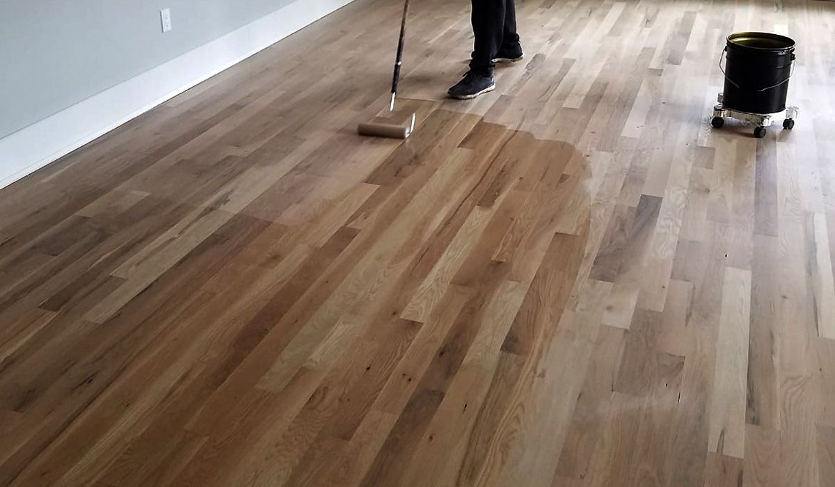 Person refinishing a hardwood floor with a mop and bucket in a mostly empty room