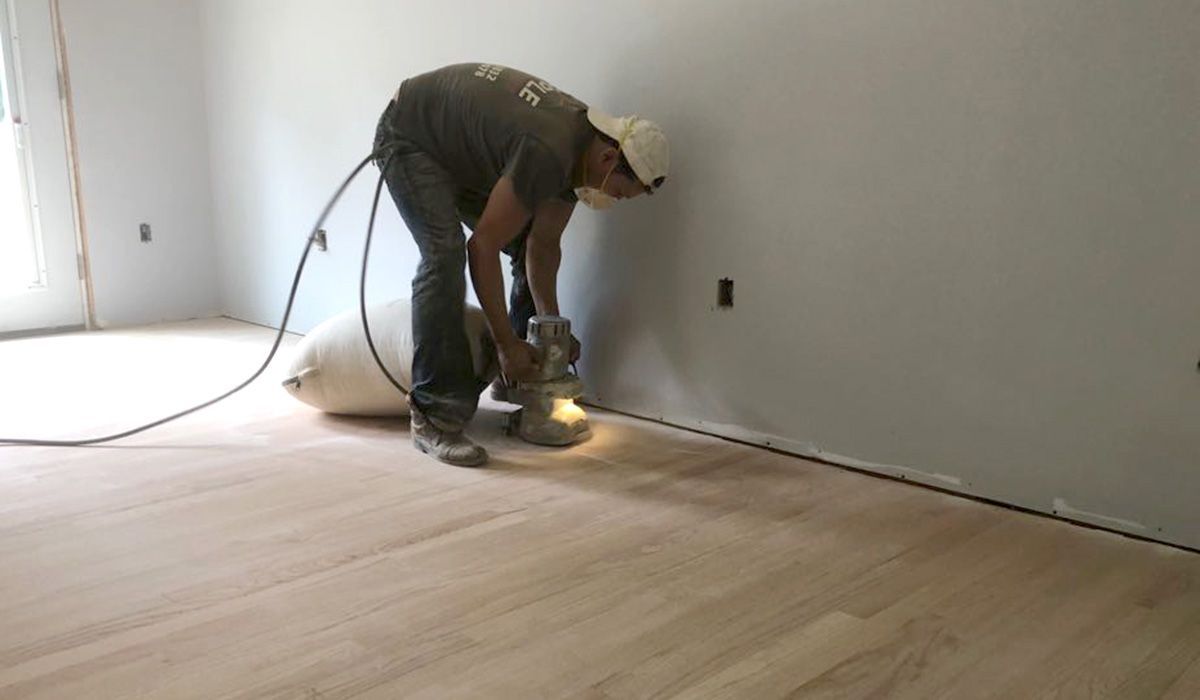 Worker sanding a hardwood floor beside a bare wall in a room under renovation