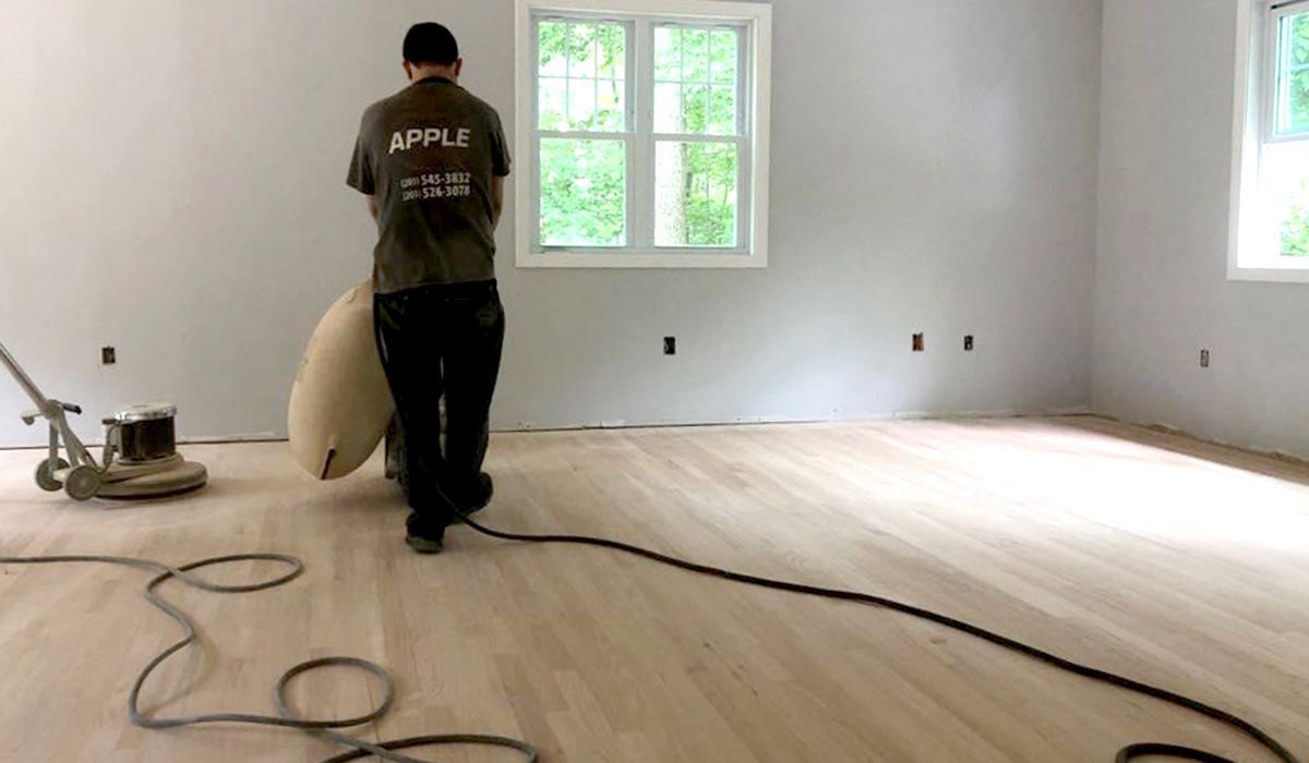 Worker sanding a light wood floor in an empty room with windows and coiled cables.