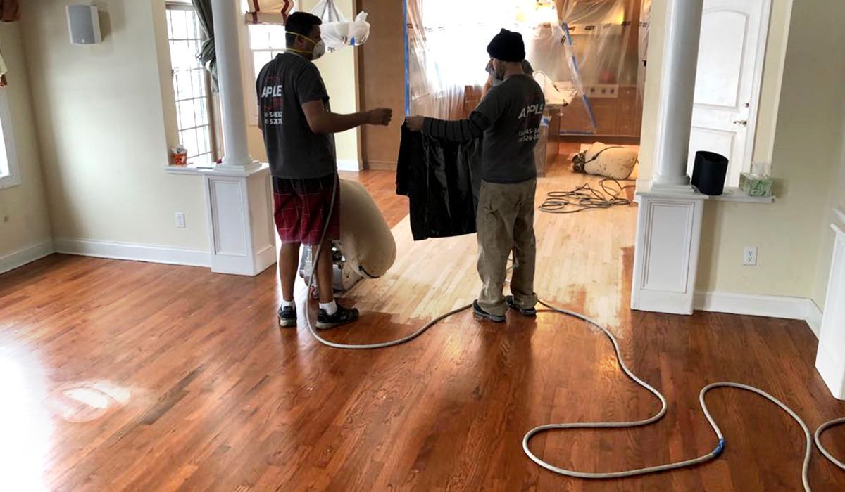 Two workers refinishing a hardwood floor in an empty room, with sanding equipment and extension cords.