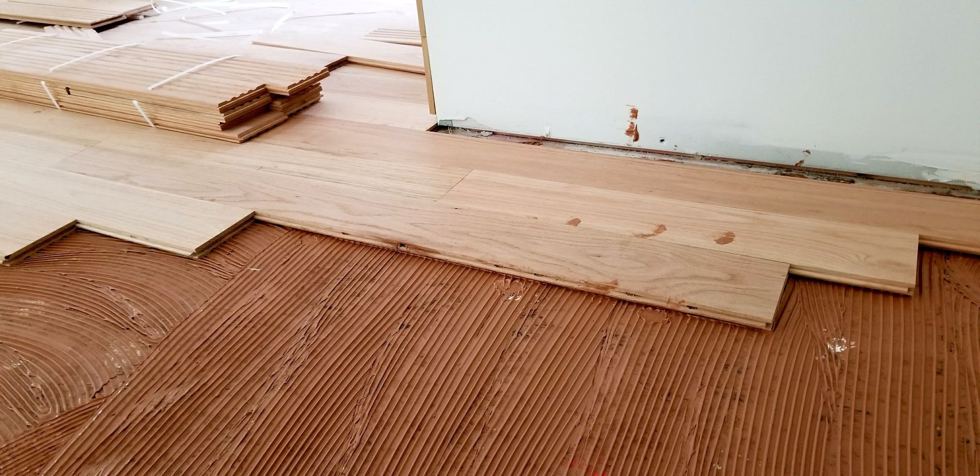 Partially installed wood flooring sheets over brown subfloor beside a white wall