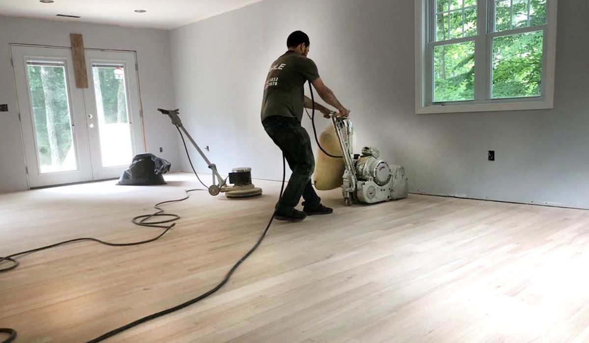 Person sanding a hardwood floor in a bright room with tools and extension cord nearby