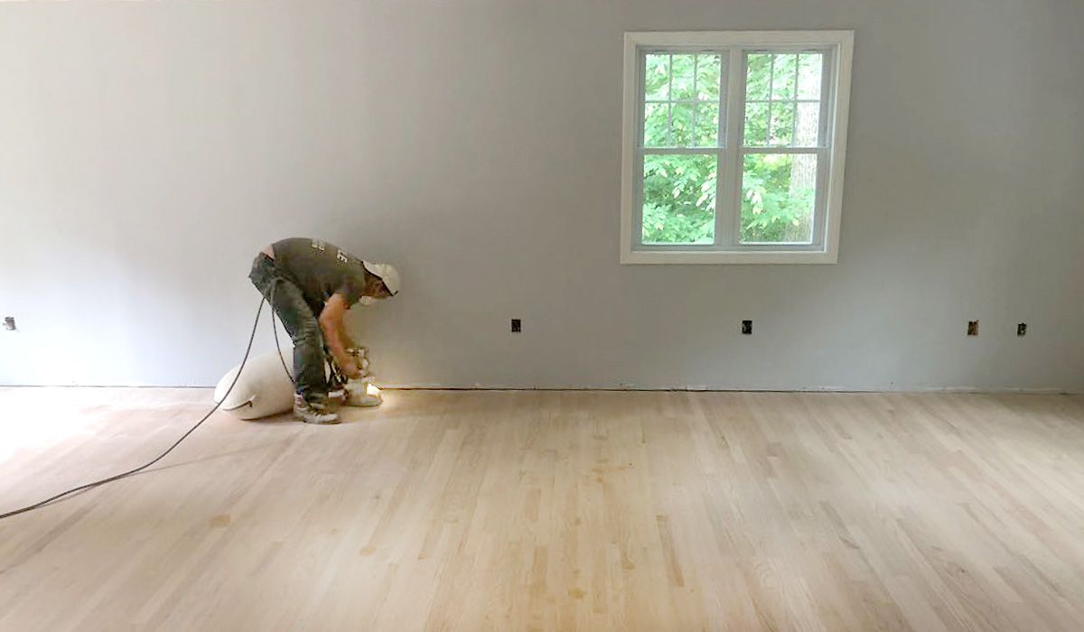 Worker sanding hardwood floor in a nearly empty room with a window and white walls