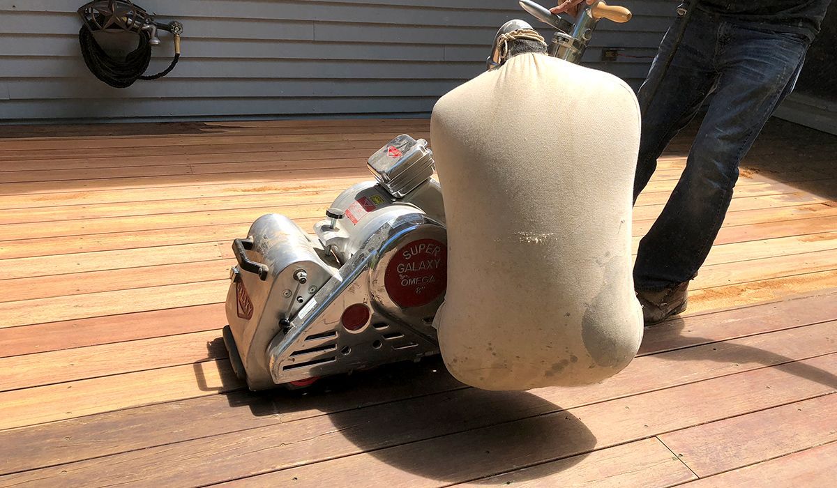 Worker using a machine to sand a wooden deck, with dust collection attached.
