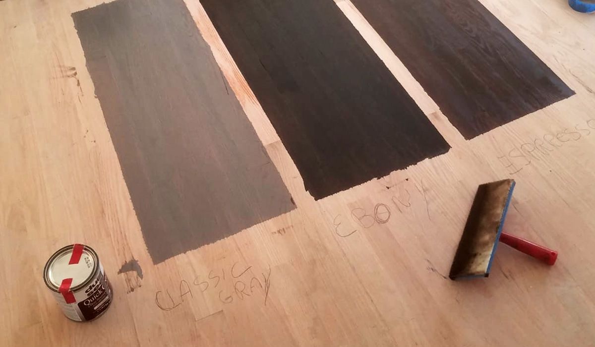 Paint roller and paint can beside three dark brown painted floor strips on a wooden floor