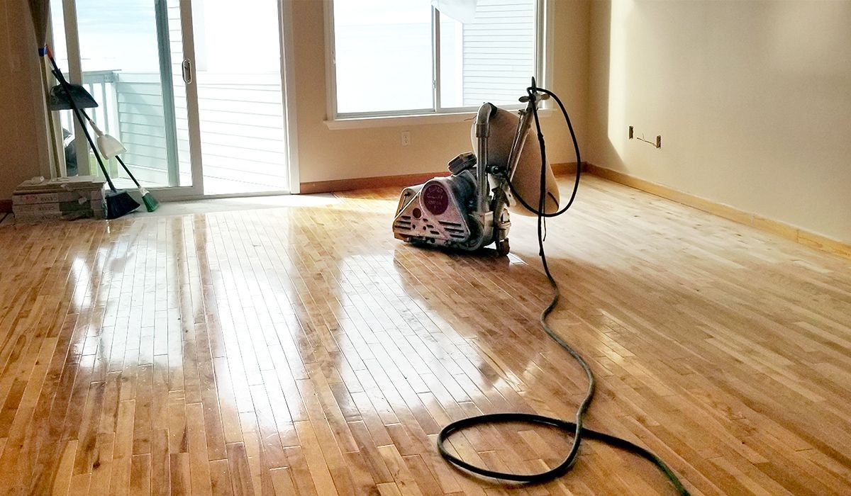 Sunlit empty room with refinished wooden floor and a floor sander near a cord by the window.