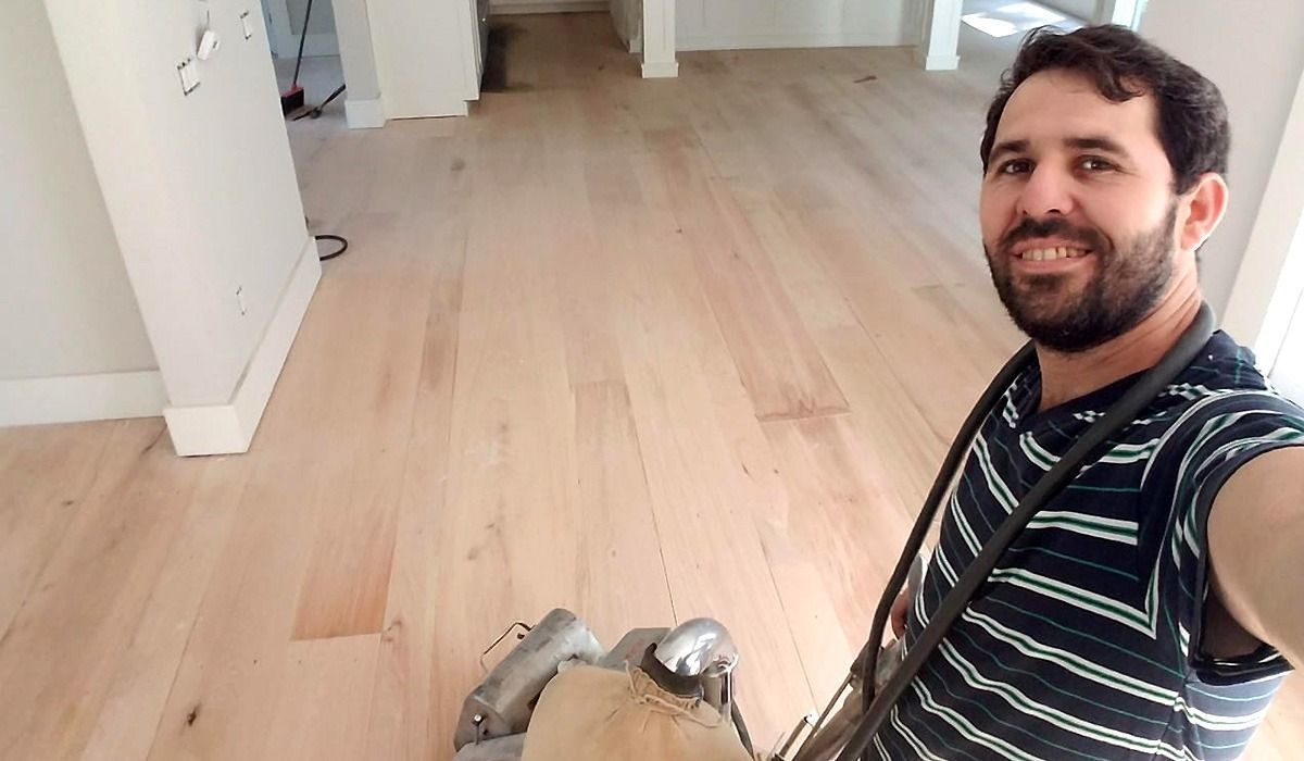 Man smiling in a striped shirt taking a selfie in an empty room with light wood floors.