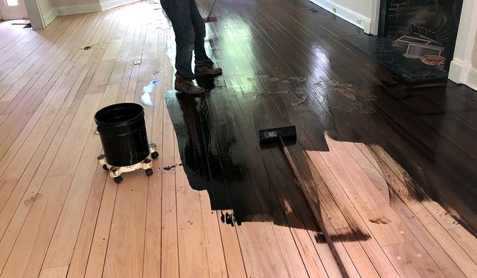Worker spreading dark stain on wooden floor with a bucket nearby in a hallway