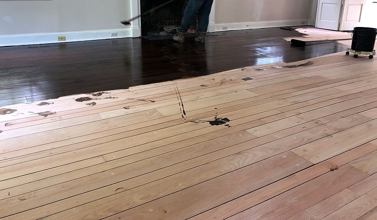 Wooden floor under construction with rows of staples, a worker in the background, and a sanding machine at right