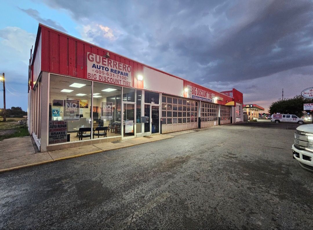 Auto repair shop at dusk with lit glass storefront and red sign along a gravel lot.