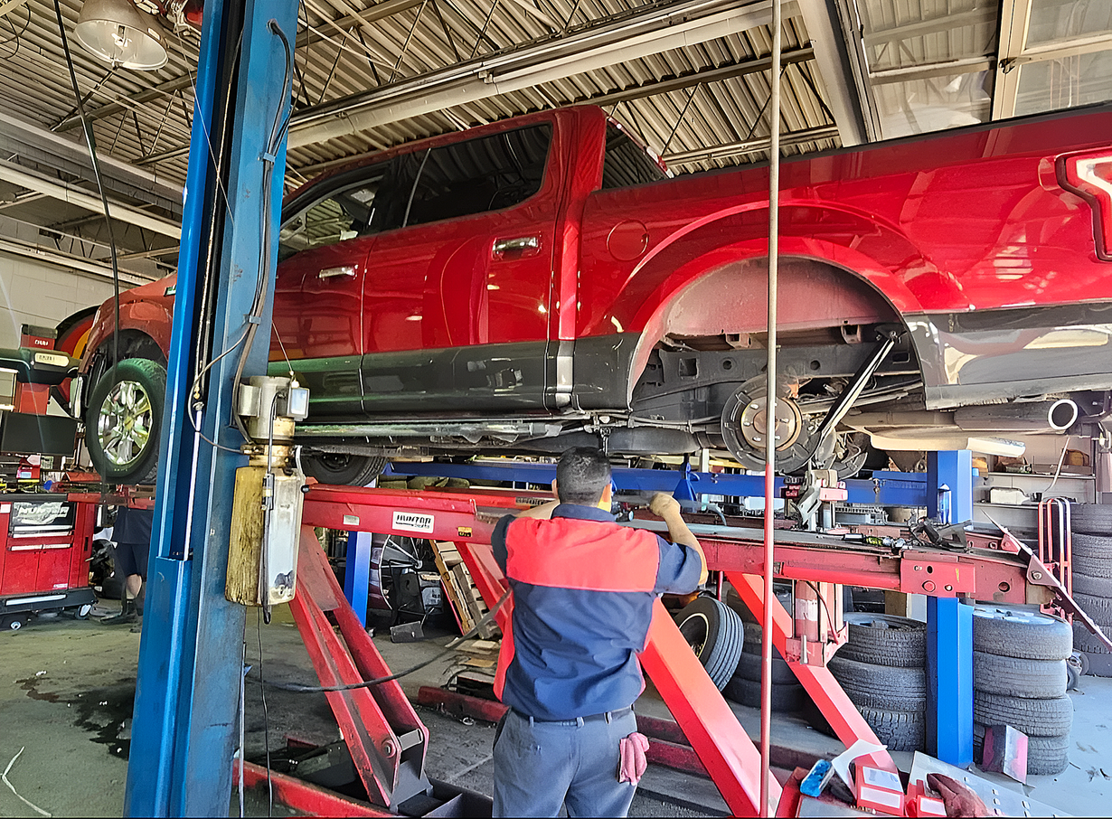 Red pickup truck on a lift in an auto repair shop, with a mechanic working underneath.