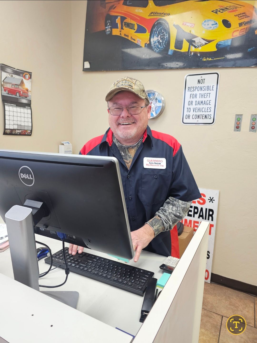 Smiling worker behind a desk in an auto shop, with a monitor and racing car poster on the wall.