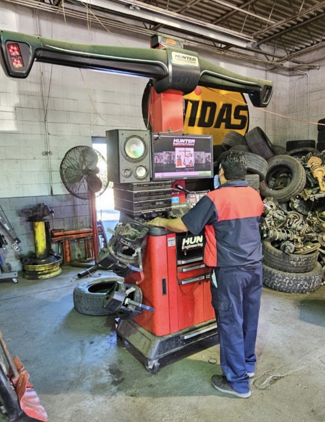 Mechanic operating red tire balancer in a workshop, with tires stacked around and a fan nearby.