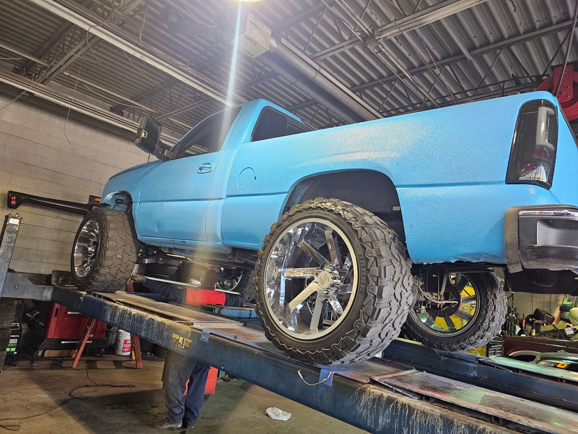 A bright blue pickup truck with large, custom wheels is raised on an automotive lift inside a repair shop.